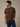 A man wearing a coffee brown short-sleeve crewneck t-shirt and blue jeans, standing against a textured gray wall. Heavyweight enough for the job, comfortable enough for after.