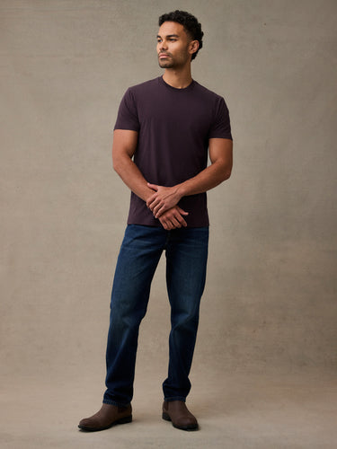 man wearing a dark plum crew-neck t-shirt, dark blue jeans, and brown boots, standing 