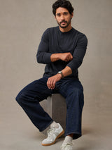 A man wearing a dark charcoal long-sleeve henley shirt, dark blue jeans, and white sneakers sits on a gray concrete block against a textured gray studio background.